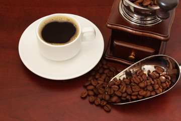 White cup of coffee, manual coffee grinder and roasted beans, metallic shovel on wooden