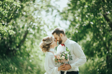 stylish bride in silk dress and groom in suit walking in sunny garden on wedding day. luxury...