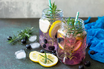 Lemonade or cocktail with blueberries and rosemary, cold refreshing drink with ice on dark concrete or stone table. Copy space.