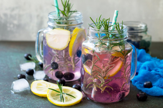 Lemonade or cocktail with blueberries and rosemary, cold refreshing drink with ice on dark concrete or stone table.