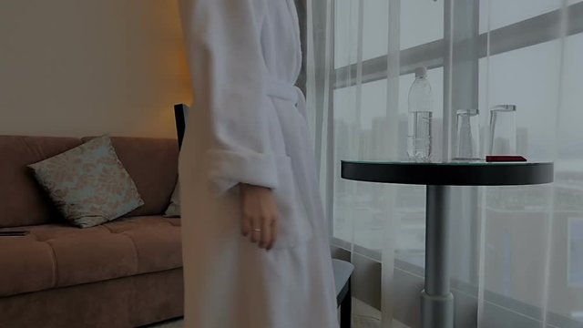 A brunette in a white robe sits at a table and drinks water at the hotel