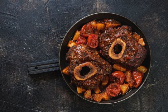 Metal pan with ossobuco made of cross cut veal shank with oranges and tomatoes. Above view on a dark brown stone background, horizontal shot with space