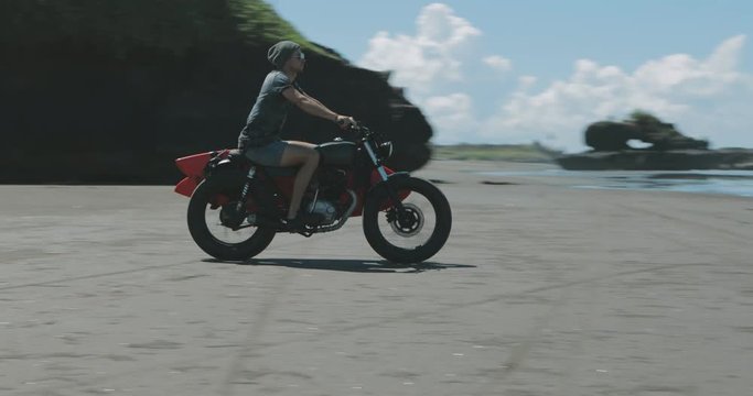 Handsome man biker surfer driving black motorbike cafe racer with red surfboard shortboard on beach to ocean at sunny day, stoping bike and looking at waves. 4k video shooting by handheld gimbal