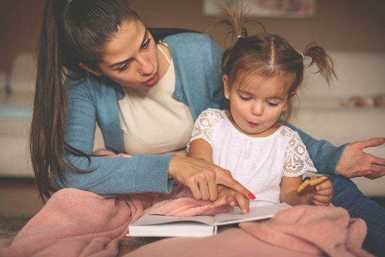 Single Mother With Her Daughter At Home Sitting On Floor And Reading Book Together.