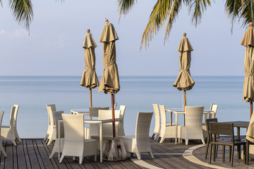White table and rattan chairs in empty cafe next to the sea water on the beach, Thailand