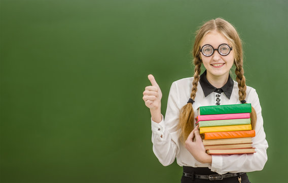 Smiling nerd student girl  with books on the background of a school board showig thumbs up. Space for text