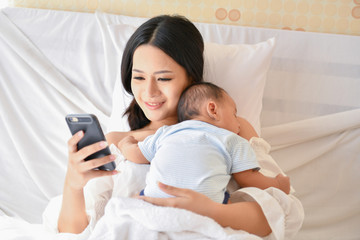 Newborn Concept. Mother and child on a white bed. Mom and baby boy playing in bedroom. Mother is playing mobile phone on bed.