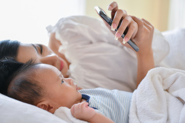 Newborn Concept. Mother and child on a white bed. Mom and baby boy playing in bedroom. Mother is playing mobile phone on bed.
