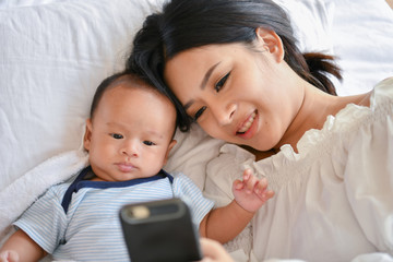 Newborn Concept. Mother and child on a white bed. Mom and baby boy playing in bedroom. Mother is playing mobile phone on bed.