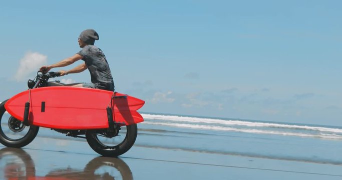 Handsome man biker surfer driving his black motorbike cafe racer with red surfboard shortboard on the beach along the ocean at sunny day. 4k video shooting by handheld gimbal