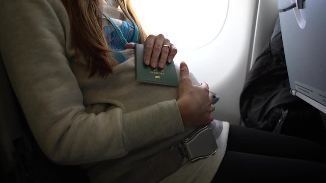 4k, Asian pregnant woman holding chinese passport in her hand and touching her tummy inside airplane. Passenger sitting on plane ready for international travel with paper identification documents-Dan