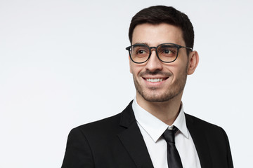 Closeup shot of young corporate advisor in trendy spectacles pictured isolated on grey background wearing formal black suit, showing toothy open smile, looking to the left, positive and confident