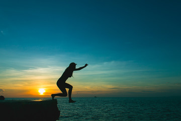 Silhouette of brave girl jumping off cliff to ocean at splendid sunset in the island of Ko Phangan, Thailand. Dare, no fear concept