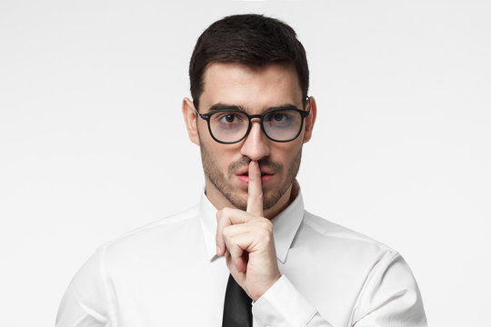 Studio picture of young business man isolated on grey background, pressing index finger to lips as if asking other to keep silent about secret or data