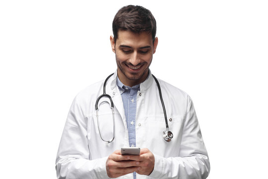 Closeup photo of man doctor standing isolated on white background looking attentively at screen of cellphone, smiling nicely