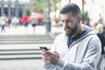 Obraz na płótnie Canvas Full bearded man using cellphone outdoors in the city. Young hipster with gray hoodie sending text message with black mobile device on street. Communication, wireless internet concept