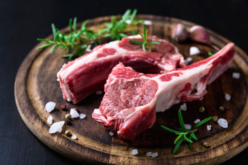 Two raw lamb chops on a wooden cutting board with spices on a dark wooden background