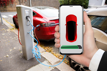 Hand with phone on a background of electric car charging point. On a device screen indicator of...