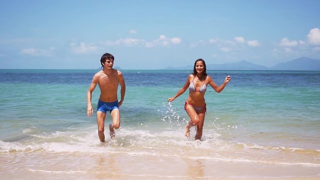 Young happy tan couple running go ut from the tropical sea in slow motion on sunny day. 1920x1080