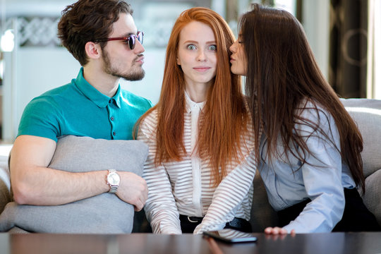 brunet young unshaven man in sun glasses and brunnet woman is going to kiss pretty red-haired woman with wide open eyes.three mates. funny precious friends