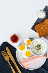 Breakfast, half boiled eggs, ham, fresh cheese sprinkled with dill, toast, horseradish sauce and sweet and sour sauce. Black kitchen cloth with golden cutlery. Flat lay on white background.