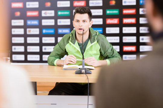 Waist-up portrait of handsome young soccer player looking at journalists and answering their questions while participating in press conference after successful completion of match