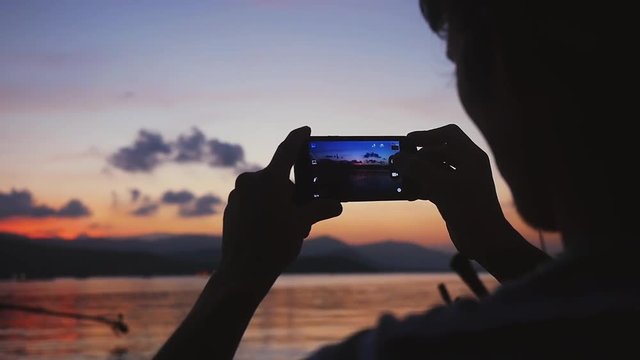 Silhouette of man standing on beach taking photo with smart phone of beautiful sunset on his vacation. slow motion. 1920x1080