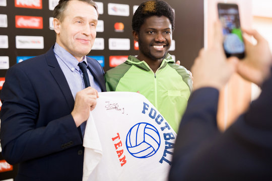 Cheerful African American football player and his middle-aged coach posing for photography with autographed T-shirt in hands while standing against press wall