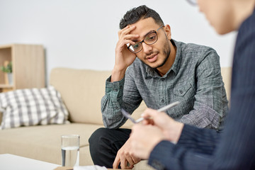 Close-up shot of young psychologist taking notes on clipboard while consulting depressed mixed-race patient during therapy session at cozy office - obrazy, fototapety, plakaty