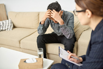 Despaired young man sitting on sofa of cozy psychotherapy office and sharing problems with highly professional psychologist at therapy session