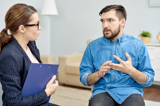 Depressed patient wearing denim shirt having face to face problem discussion with highly professional psychologist after completion of group therapy session, interior of cozy office on background