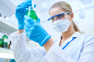Serious confident young female scientist wearing protective goggles and mask learning hazardous...