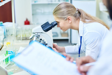 Concentrated young female biologist looking through microscope while analyzing sample in modern...