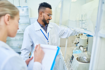 Positive content confident young Arabian scientist in lab coat adjusting laboratory equipment and reporting medical assistant about results