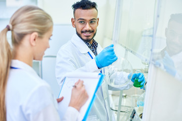Cheerful skilled handsome young Arabian male chemist with beard holding laboratory glassware and...