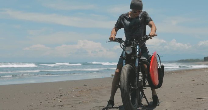 Handsome man biker surfer sitting down on black motorcycle cafe racer with red surfboard shortboard on beach at sunny day and turning engine. 4k video shooting by handheld gimbal