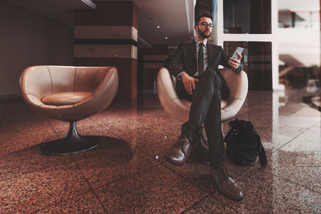 Young successful man entrepreneur with beard, in formal suit and glasses is sitting in armchair next to another empty one in reception area of his office, using cellphone while waiting for a meeting