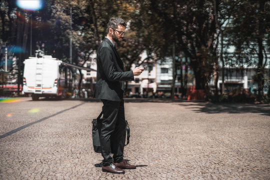 Young handsome bearded businessman in a formal suit and glasses is standing on a pavement stone outdoors, calling taxi via an app on his smartphone; with copy space place for advertising or your text