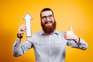 Cheerful bearded man pointing up with arrow and wearing eyeglasses and showing thumbs up