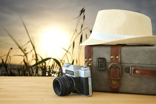 traveler vintage luggage, camera and fedora hat over wooden table infront of a field at sunset light. holiday and vacation concept.