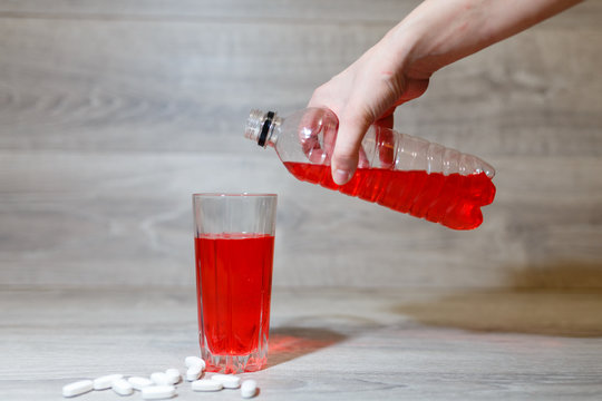 A Woman's Hand Pours A Red Sports Drink Or Lemonade Into A Glass Cup From A Plastic Bottle. Energy Drink In Glass And Plastic Bottle On Wooden Table, Scattered Pills. Energy For Workout In A Gym.