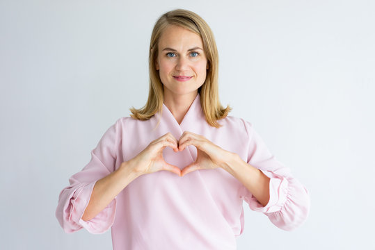 Smiling Peace-loving Lady Showing Heart And Looking At Camera. Positive Attractive Woman In Pink Blouse Expressing Kind Relation. Mercy Concept