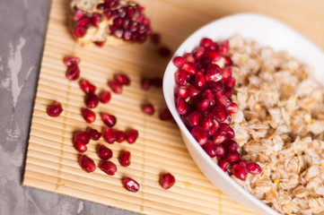 Cooked yummy warm oatmeal and heap of ripe fresh pomegranate seeds in white ceramic bowl near piece of pomegranate on clean bamboo rug on worn gray scratched concrete with copy space
