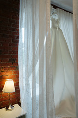 The bride's dress in the hotel room hangs over the bed in anticipation of the mistress. The table lamp burns
