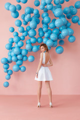 Young woman in white cocktail dress standing back to camera on pink wall background with blue bubbles hanging. Dreaming concept.