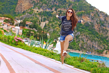 young happy beautiful woman in glasses, shorts and sneakers jumping in the resort near the nature, sea, mountains and flowers, the girl laughs and smiles