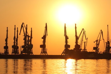 Obraz premium Seaport at sunset, cranes in the port on the horizon, orange sky against the sea horizon, landscape with high-rise cranes, construction of a breakwater in the sea