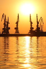 Fototapeta na wymiar Seaport at sunset, cranes in the port on the horizon, orange sky against the sea horizon, landscape with high-rise cranes, construction of a breakwater in the sea