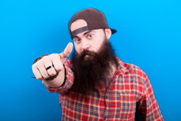 Naklejka premium Long bearded man showing thumbs up to the camera and only the hand is in focus. Blue background in studio photo