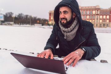 A young guy with a beard working on a laptop in the winter on the street is very cold computer all in the snow